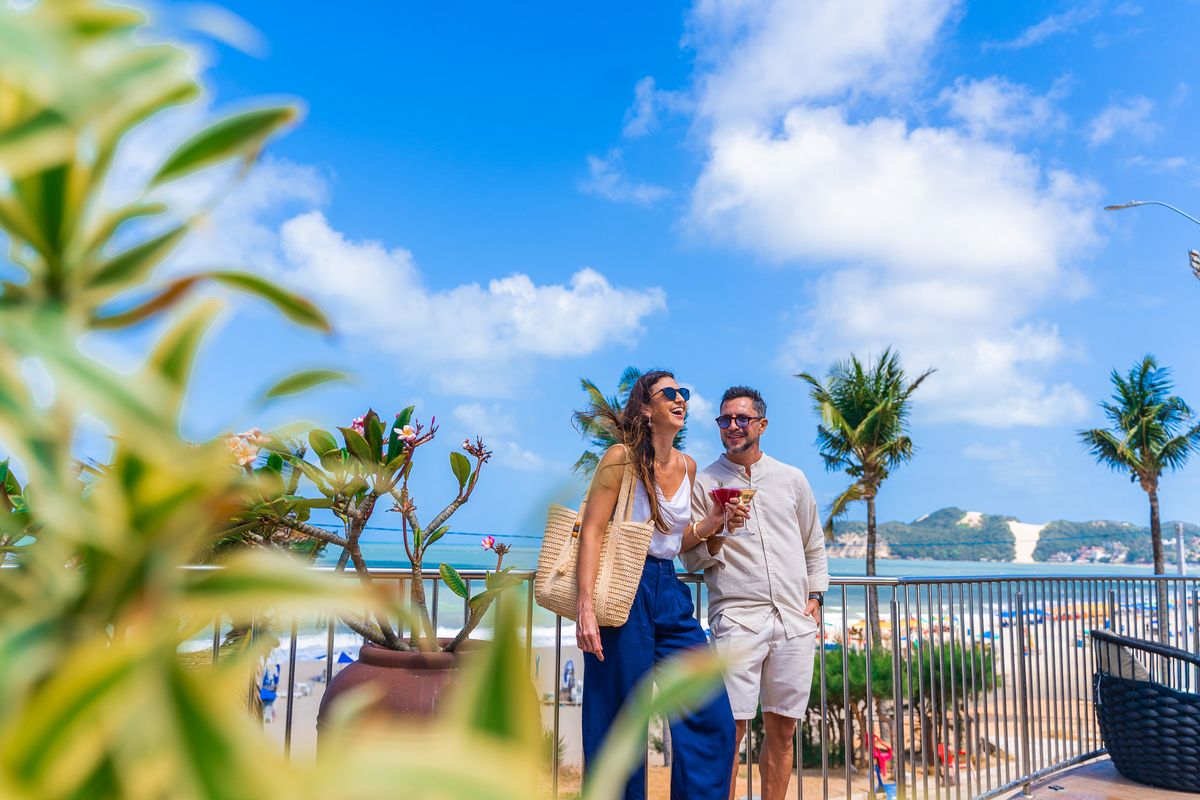 Casal sorri no deck suspenso do Esmeralda Praia Hotel com vista para o mar e o Morro do Careca
